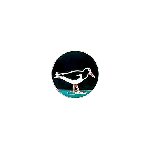 Black round enamel pin with illustrated black and white bird with orange legs and beak.